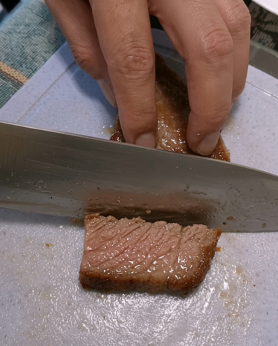 Hände beim Aufschneiden eines gebratenen Steaks auf einem Kunststoff-Schneidebrett.