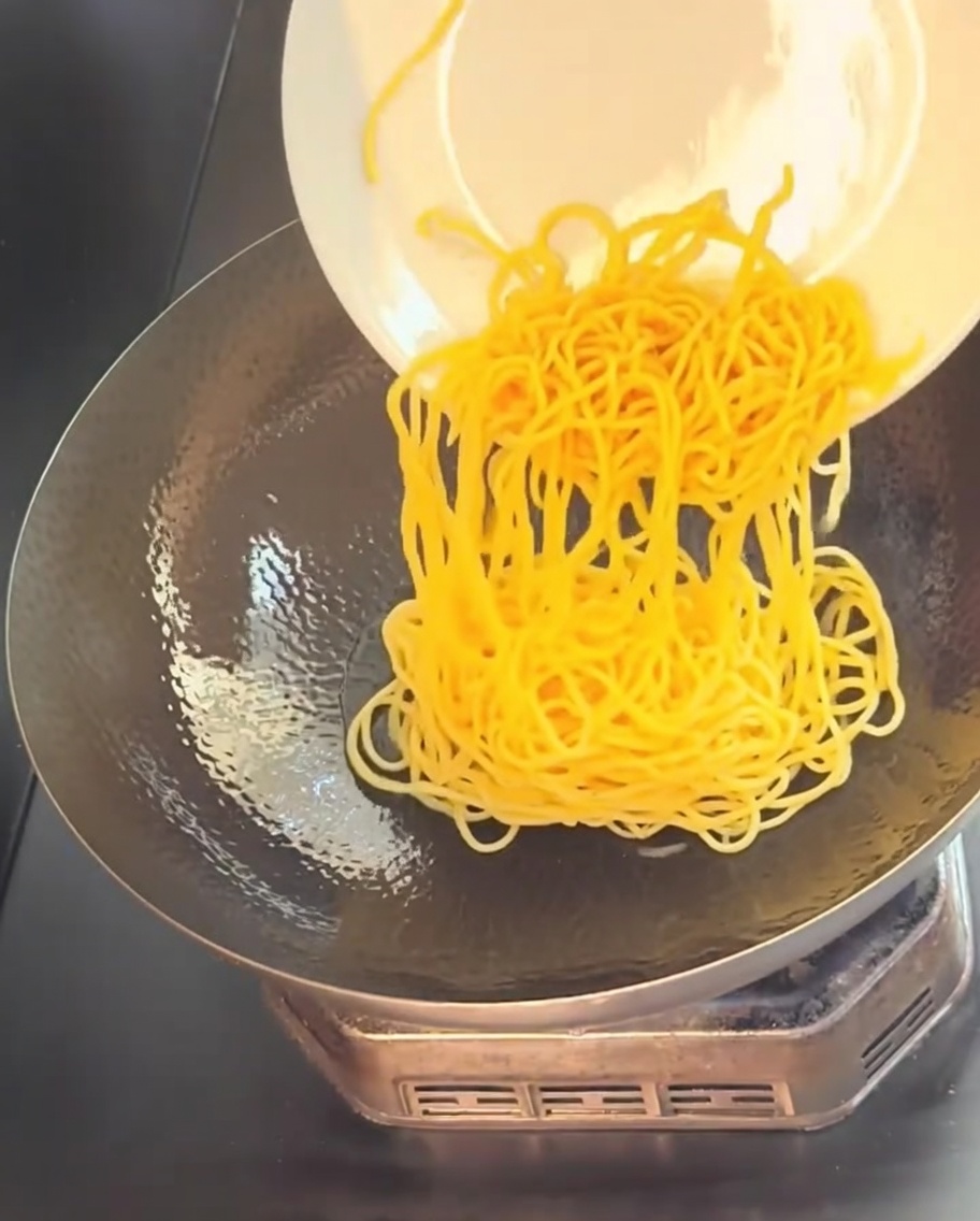 Gekochte gelbe Spaghetti werden von einem weißen Teller in einen heißen, geölten Wok geschoben.