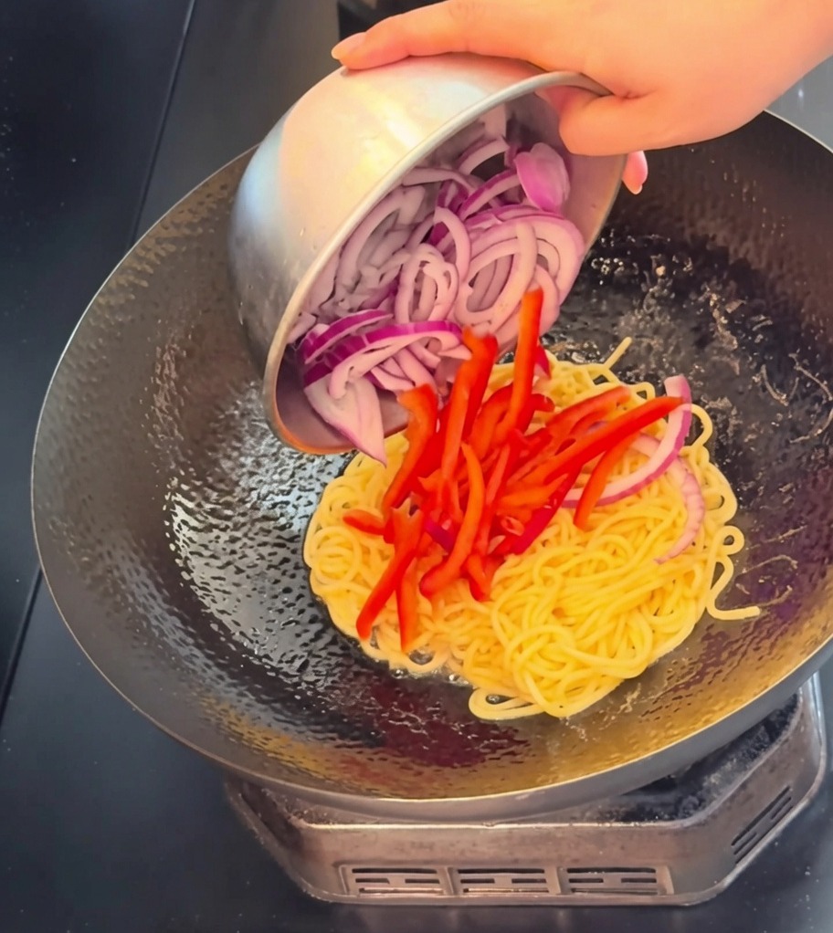 Nahaufnahme von Rindfleischstreifen, Paprikastreifen und roten Zwiebeln, die in den Wok zu den Spaghetti gegeben werden.