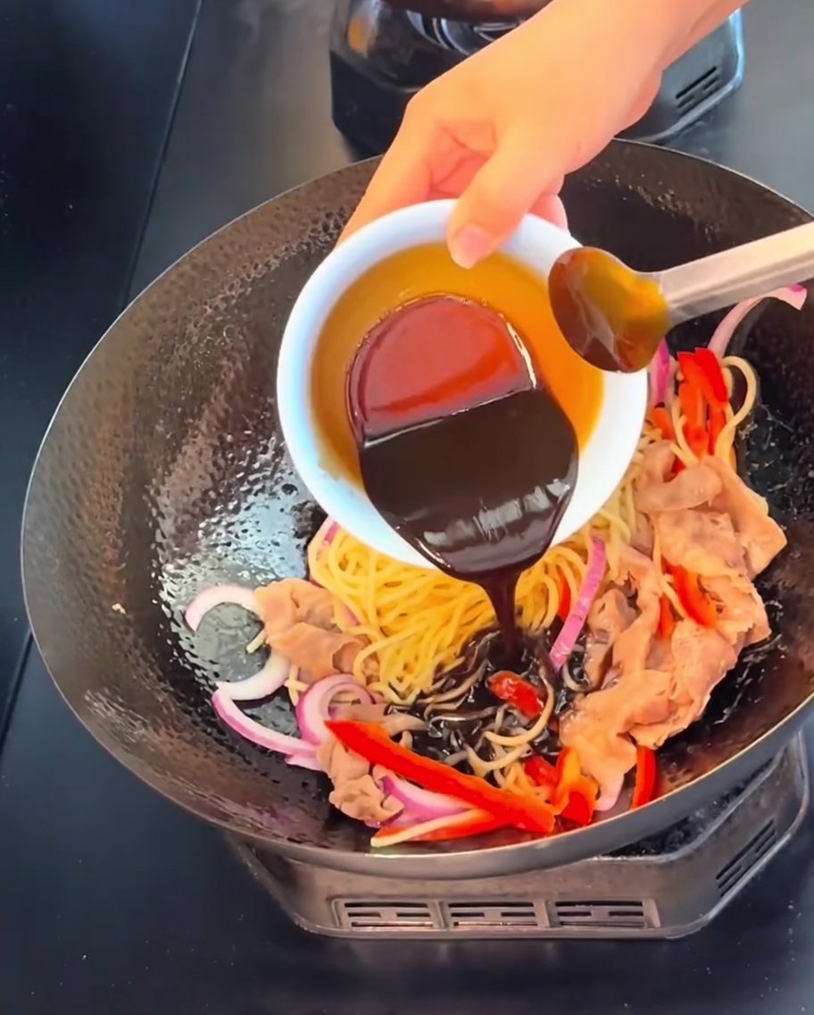 Eine dunkle, kräftige schwarze Pfeffersauce wird aus einer kleinen weißen Schüssel über Nudeln, Fleisch und Gemüse gegossen.