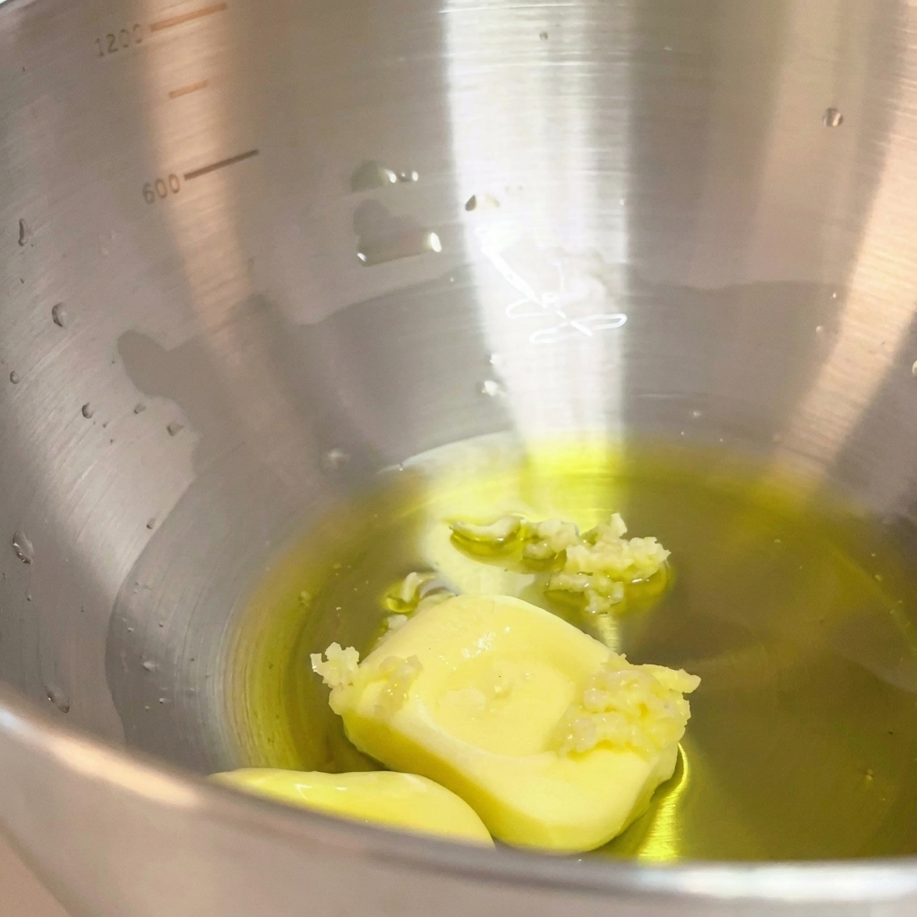 Zerdrückter Knoblauch wird zu der Mischung aus Olivenöl und Butter in der Metallschüssel gegeben.