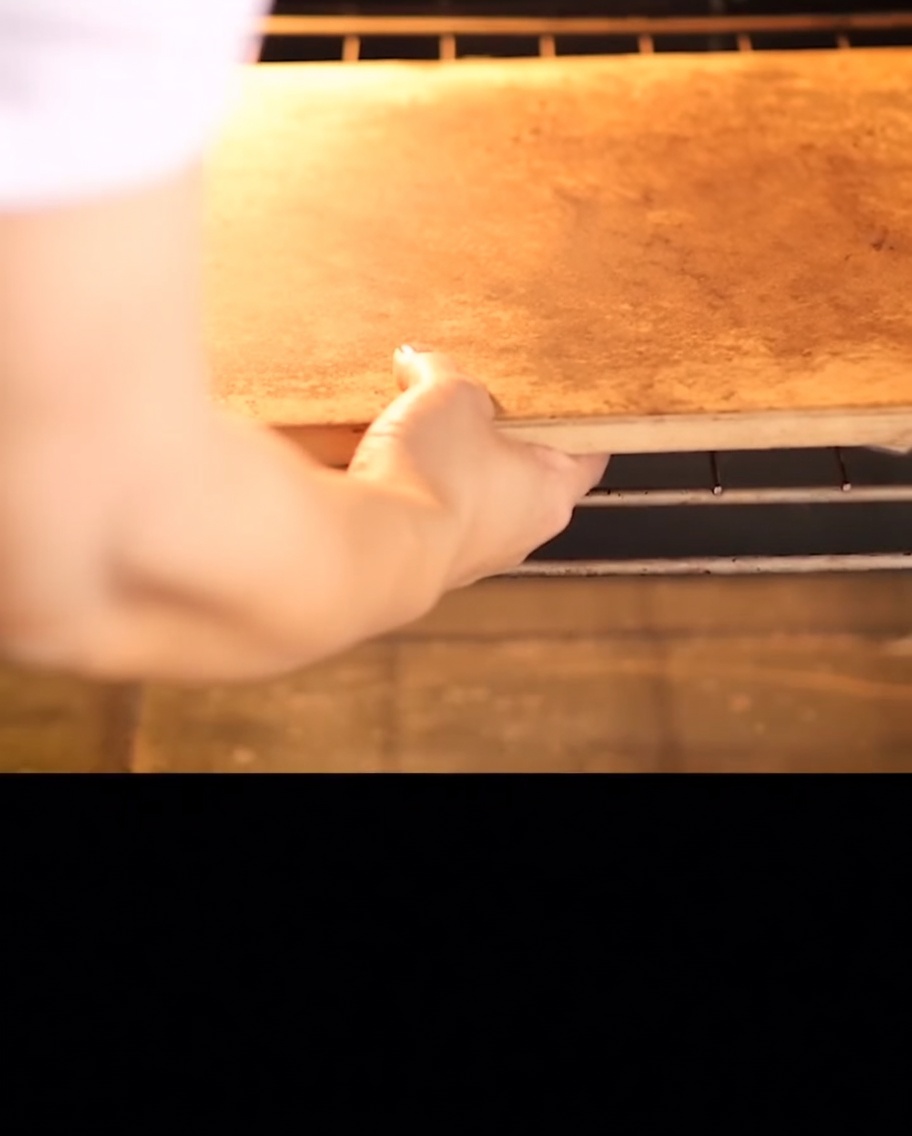 Hände schieben einen rechteckigen Pizzastein auf ein Metallgitter in einen Backofen.