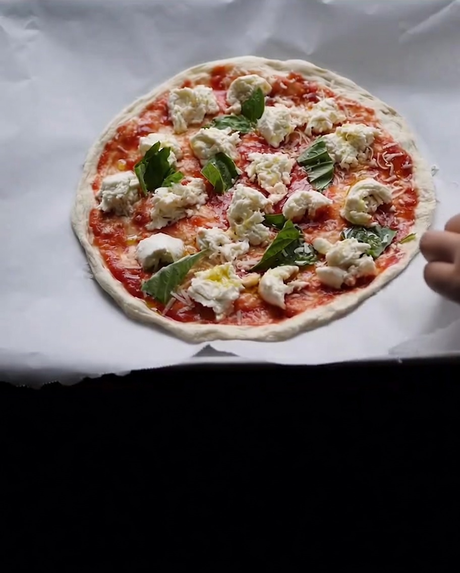 Eine rohe, belegte Pizza mit roter Sauce, frischem Mozzarella und Basilikumblättern auf Backpapier.