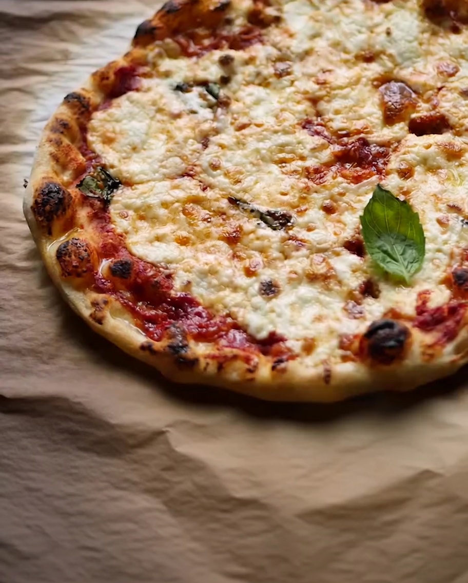 Eine frisch gebackene Pizza auf Backpapier, garniert mit frischen Basilikumblättern und dunklen, verkohlten Stellen am Rand.