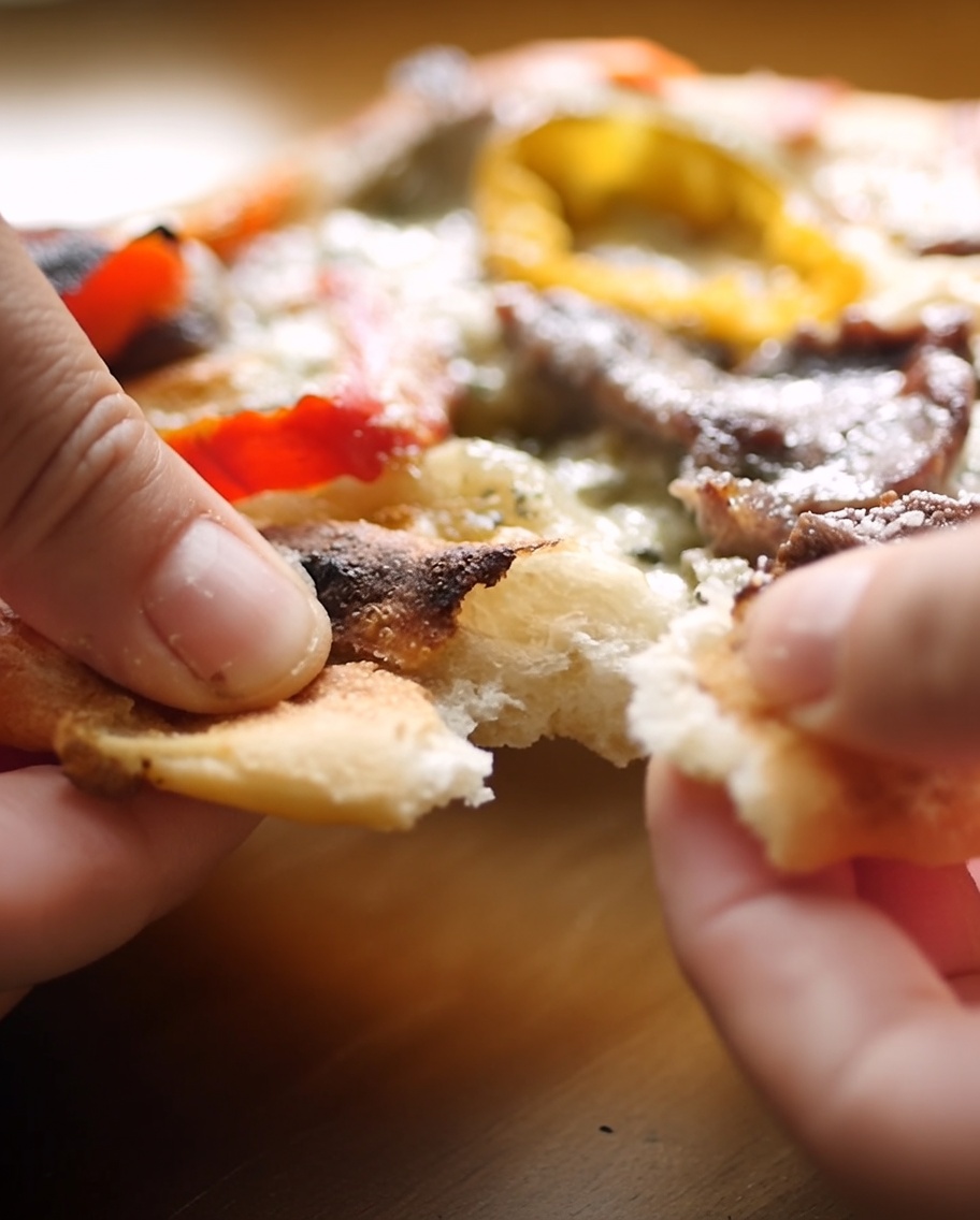 Hände reißen den Rand eines gebackenen Pizzateigs auf, um das weiche, luftige Innere und das knusprige Äußere zu zeigen.