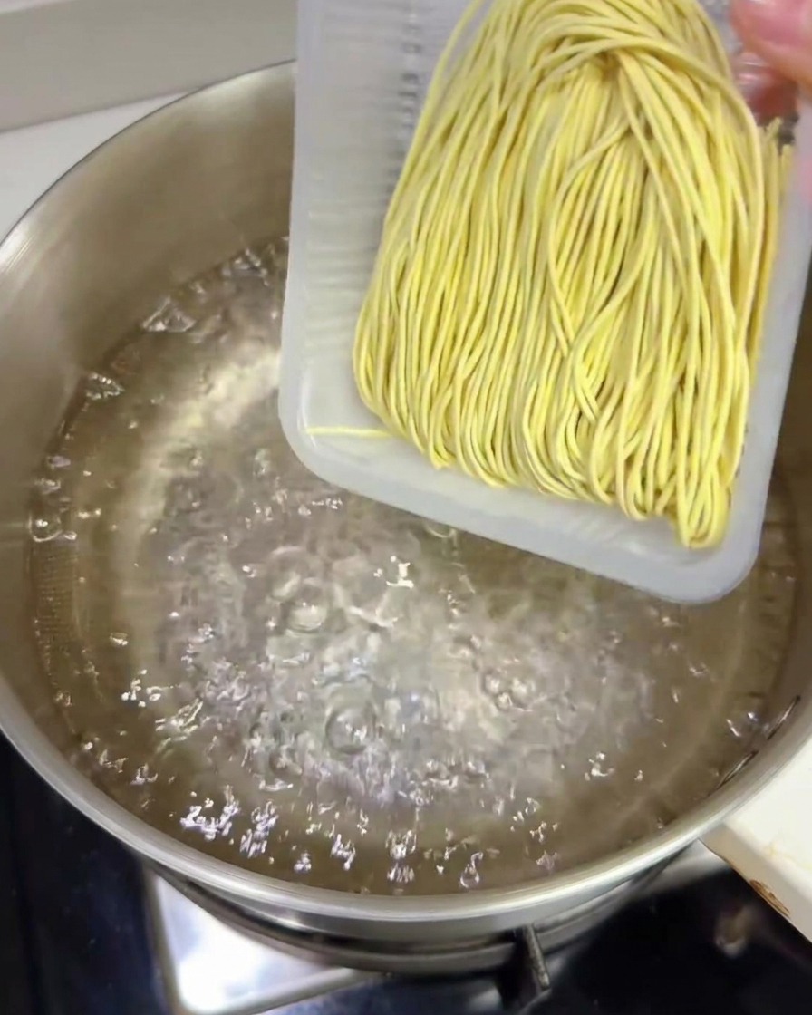 Ein Block frischer gelber Ramen-Nudeln gleitet aus einer Kunststoffschale in einen Topf mit kochendem Wasser.