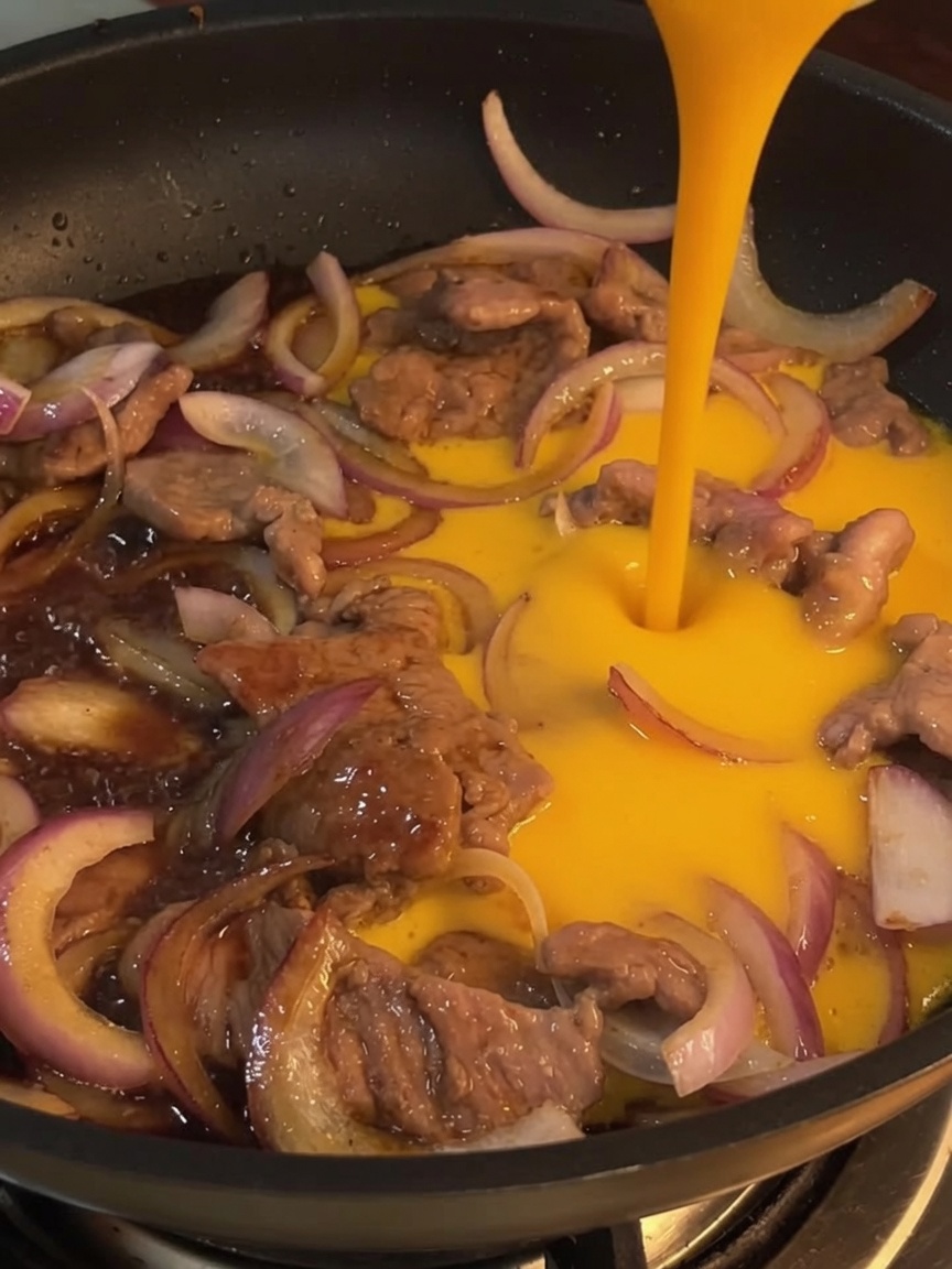 Nahaufnahme von verquirlten Eiern, die in eine Pfanne über Rindfleisch und Zwiebeln gegossen werden.
