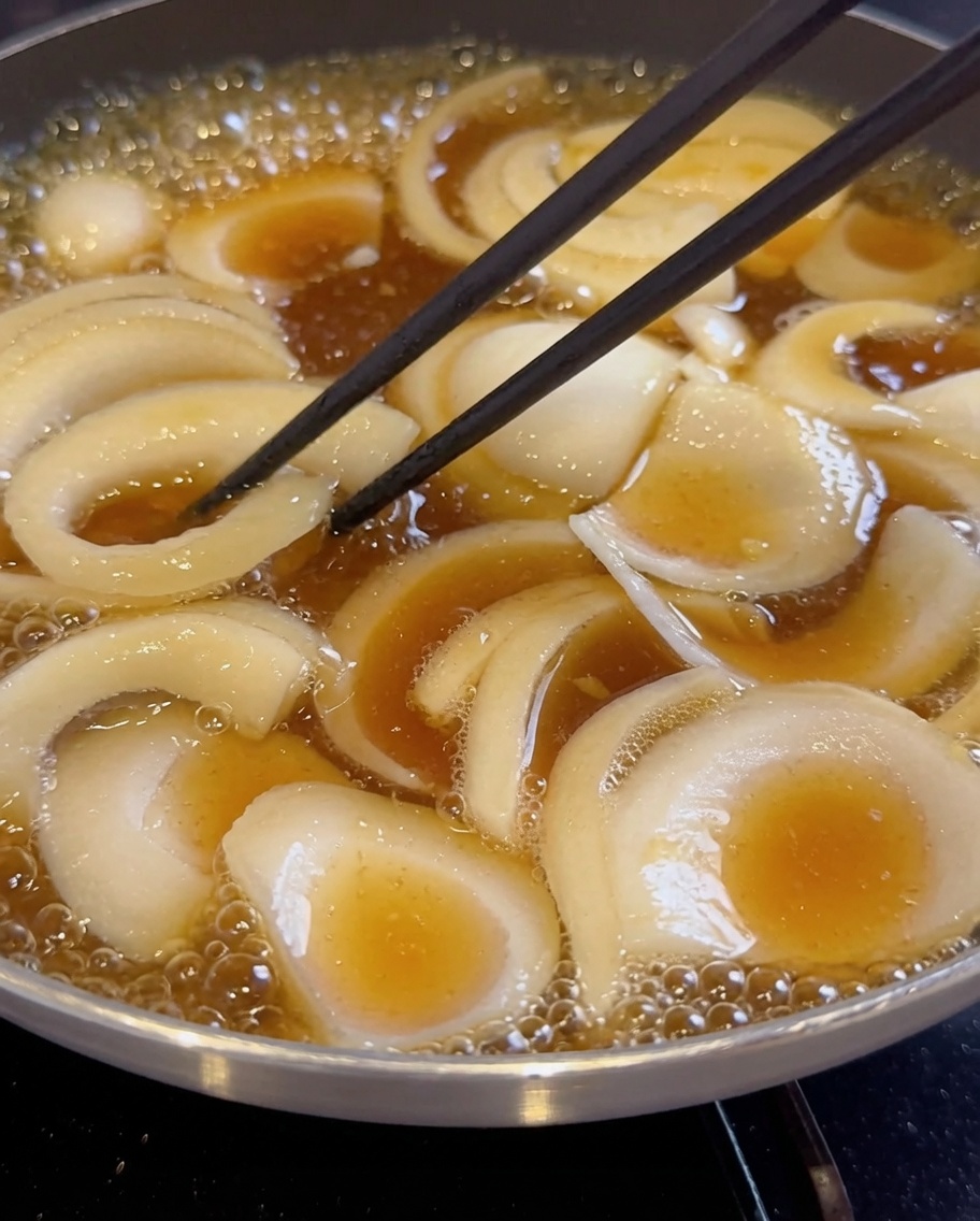 Geschnittene Zwiebeln schmoren in einer dunklen, kochenden Flüssigkeit in einer Pfanne.