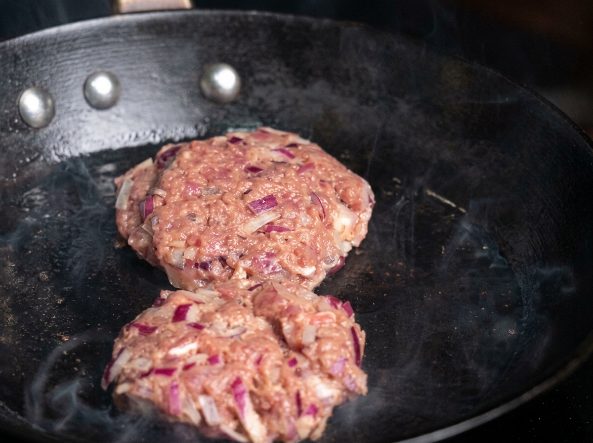 Zwei rohe Rinder-Patties werden in eine heiße, geölte Pfanne gegeben.