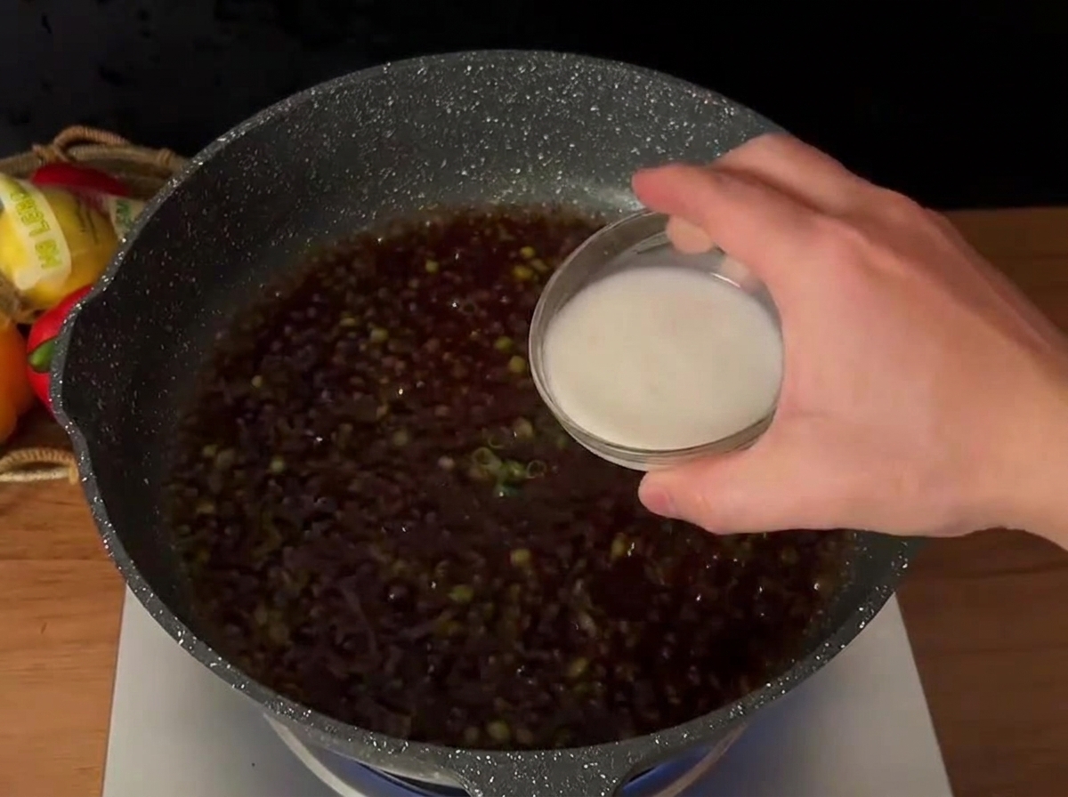 Eine Hand gießt Stärkemischung in eine kochende dunkle Sauce mit Frühlingszwiebeln.