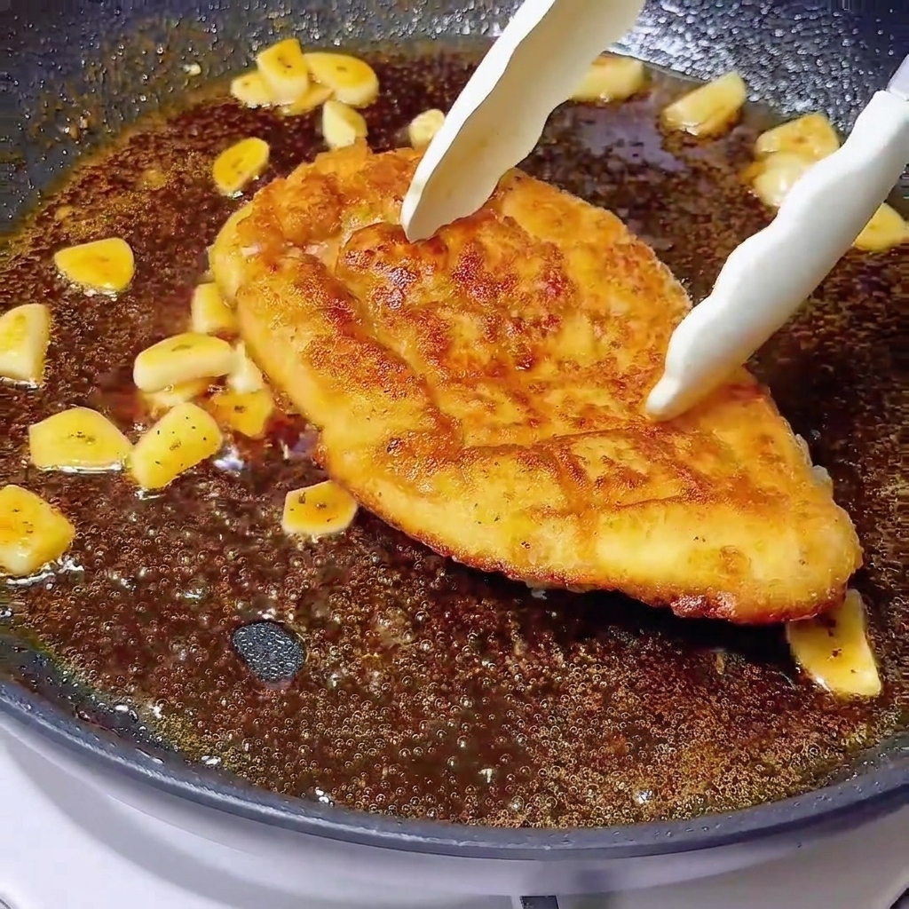 Eine Küchenzange legt eine goldbraune Hähnchenbrust in eine blubbernde dunkle Glasur mit Knoblauchscheiben.