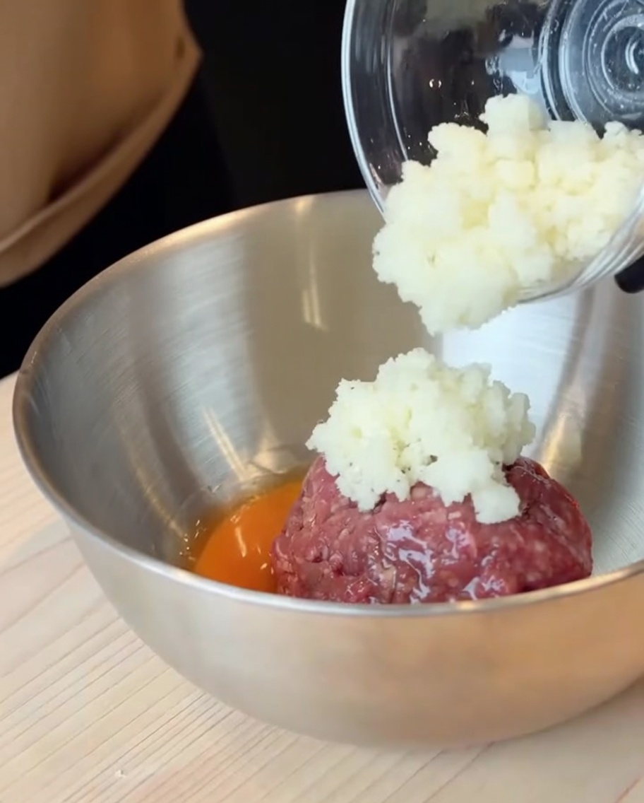 Rohes Rinderhackfleisch und ein aufgeschlagenes Ei in einer großen Edelstahlschüssel, zu der gewürfelte Zwiebeln hinzugefügt werden.