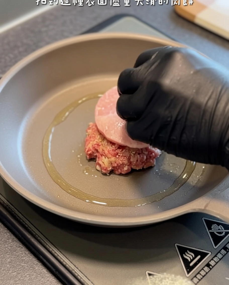 Ein rohes Hamburger Patty wird mit einem Pfannenwender und einer Hand in einem schwarzen Handschuh vorsichtig in eine heiße, geölte Pfanne gelegt.