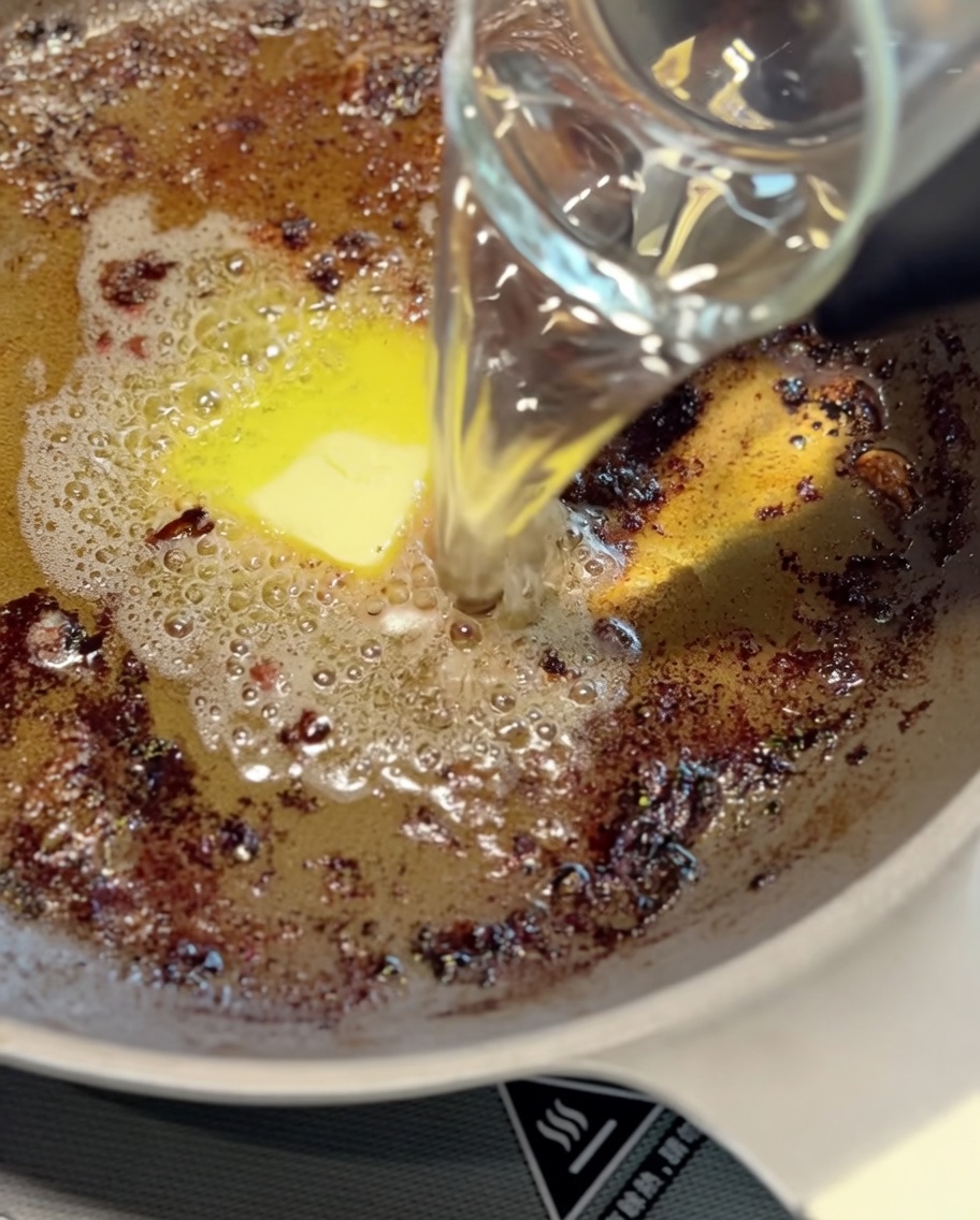 Eine Metallkelle rührt den angebratenen Bratensaft und Wasser in einer Pfanne um, um die Basis für die Soße zu bilden.