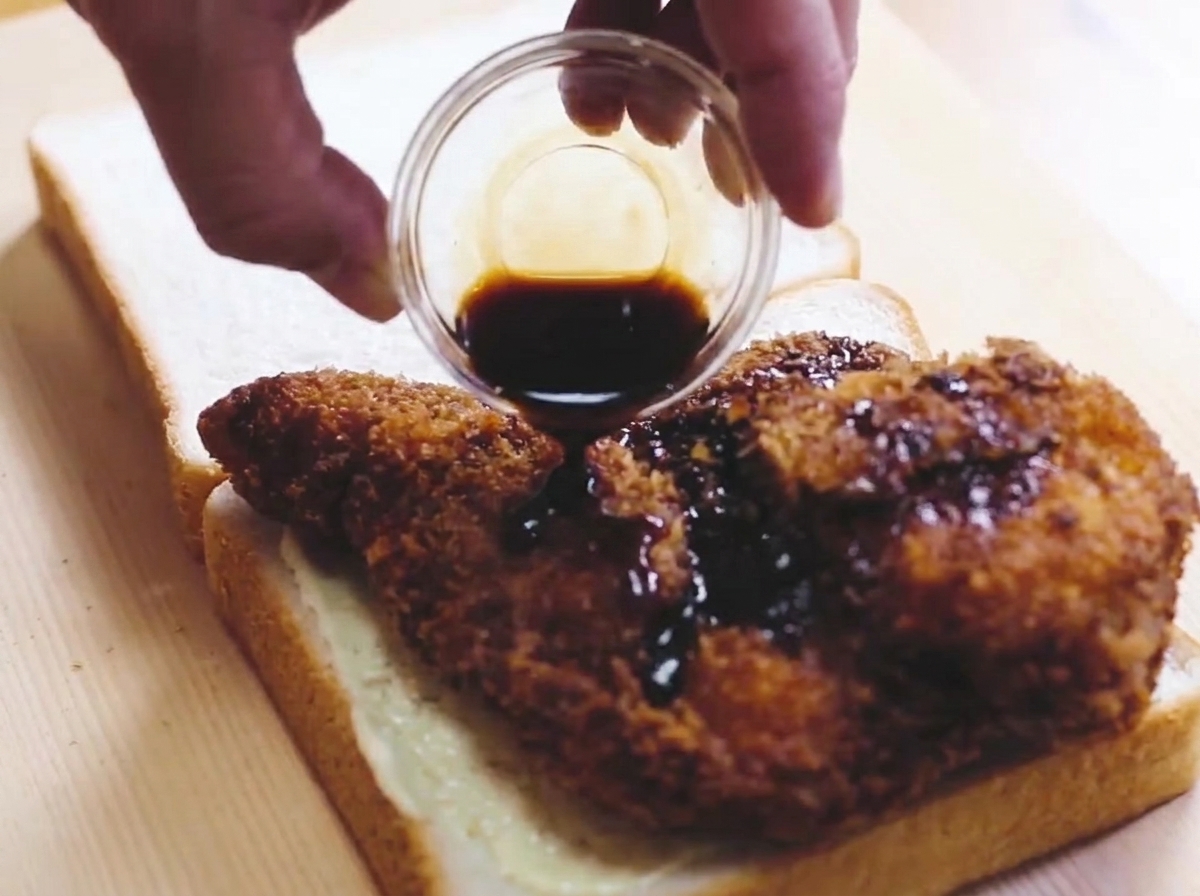Ein goldbraunes Katsu-Filet liegt auf einer Brotscheibe und wird mit Sauce beträufelt.