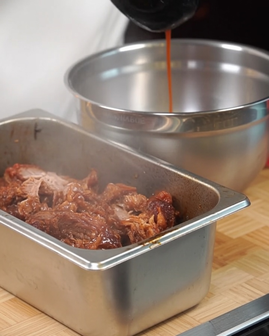 Das gezupfte Fleisch in einer Metallschale wird mit Bratensaft beträufelt.
