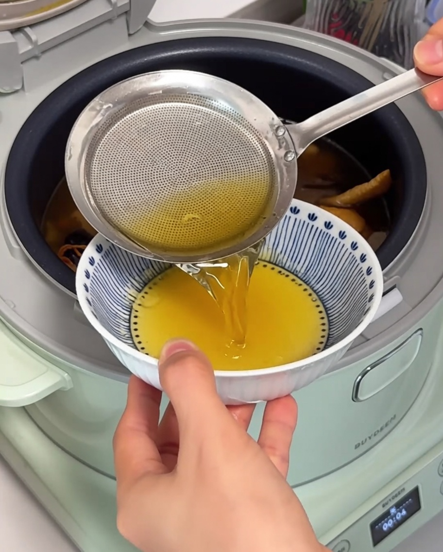 Ein feinmaschiges Sieb hebt goldenes Hühnerfett von der Oberfläche der köchelnden Suppe ab.