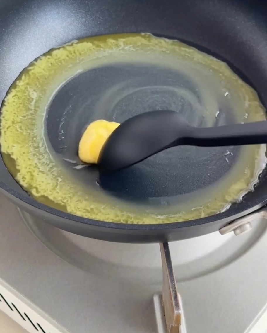 Eine dunkle beschichtete Pfanne mit geschmolzener Butter und Mehl, das mit einem schwarzen Silikonspatel zu einer Mehlschwitze verrührt wird.