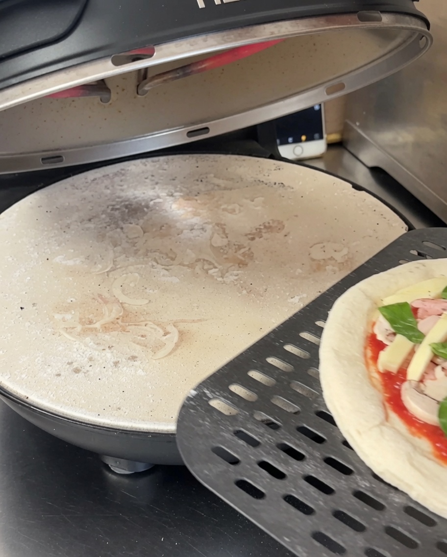 Ausgerollter Pizzateig liegt auf einem schwarzen, perforierten Pizzastein in einem Pizzaofen.
