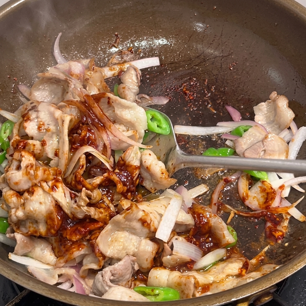 Scharfer Schweinebauch wird in der Pfanne mit einem Spatel gut mit der Sauce vermengt.