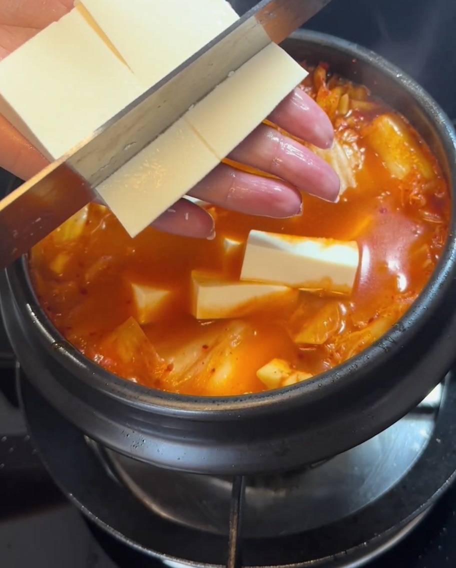 Tofuscheiben werden vorsichtig in den sprudelnden roten Kimchi-Eintopf gelegt.