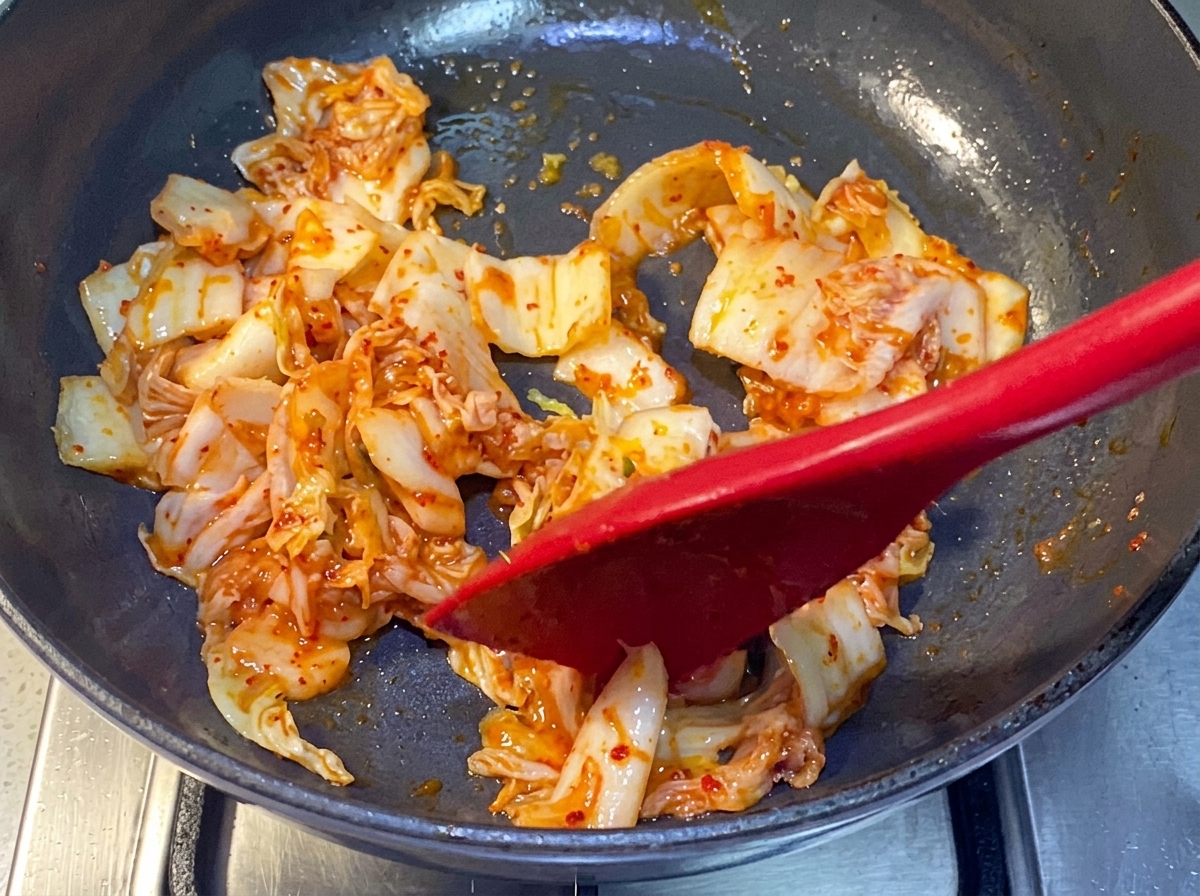 Kimchi wird mit einem roten Silikonspatel in einer Pfanne angebraten.