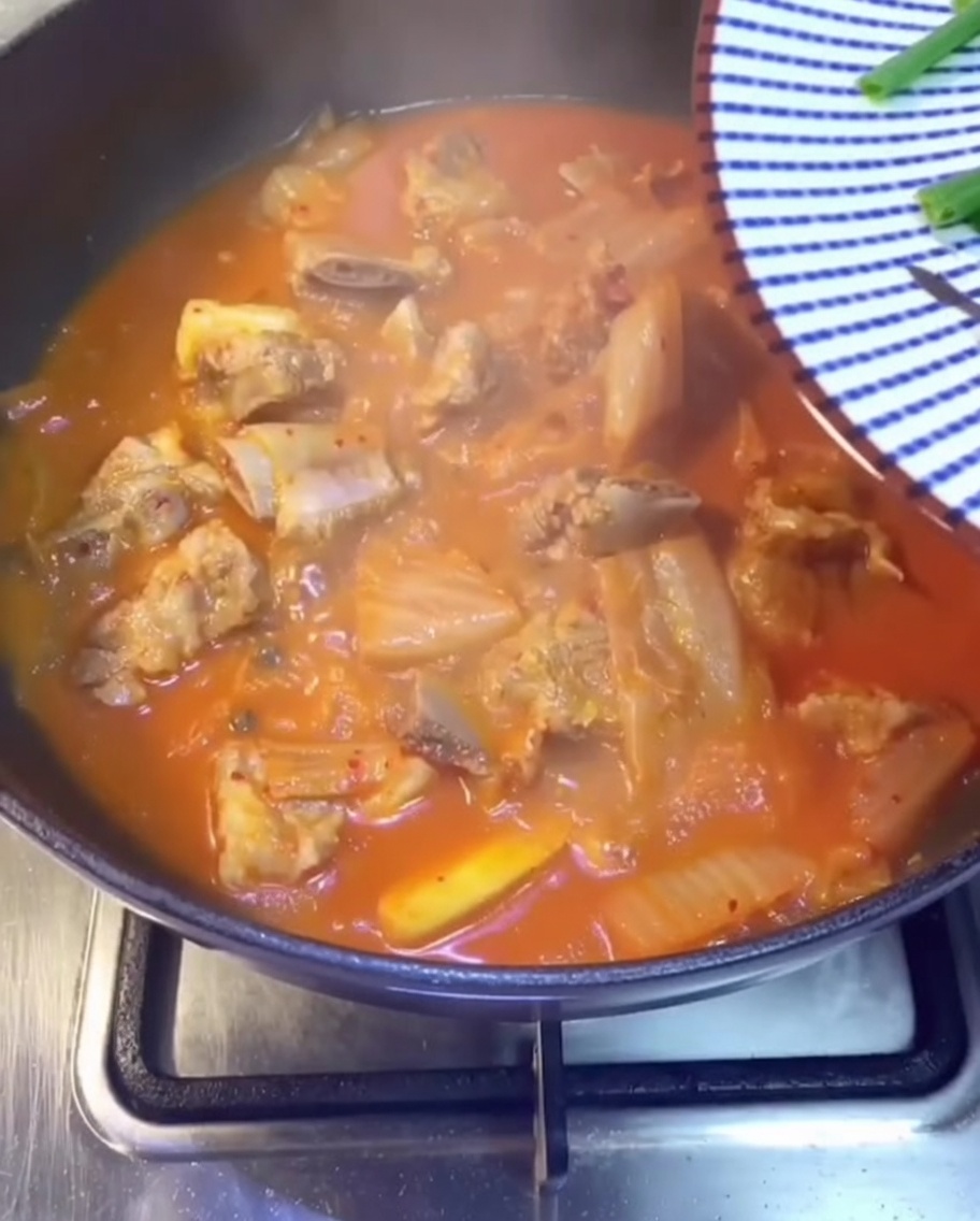 Eine Nahaufnahme von frischen Frühlingszwiebeln, Koriander und in Scheiben geschnittenen grünen Chilischoten, die über den blubbernden roten Kimchi-Schweinerippchen-Eintopf gestreut werden.