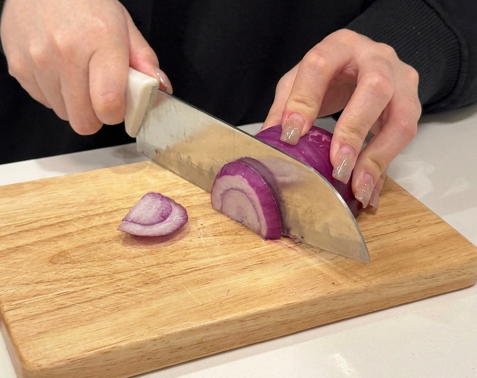 Hände beim Schneiden einer roten Zwiebel auf einem Holzbrett.
