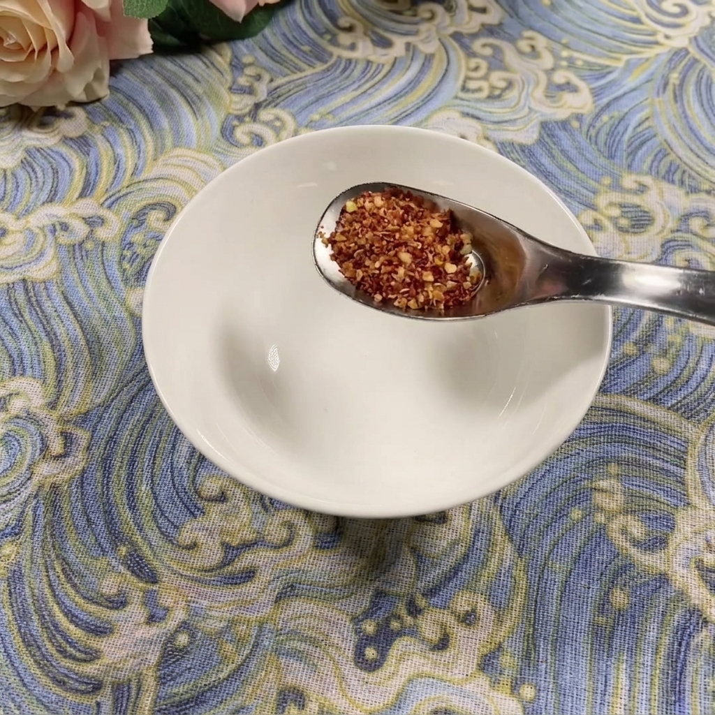 Ein Löffel grobes Chilipulver wird über einer leeren weißen Schüssel auf einer blau gemusterten Oberfläche gehalten.