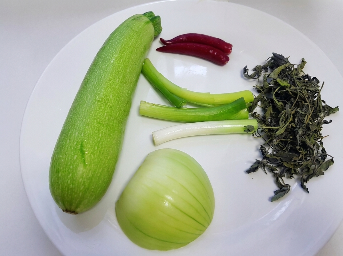 Rohe Zutaten wie ein ganzer grüner Squash, Frühlingszwiebeln, weiße Zwiebel, wilde Perillablätter und rote Chilischoten liegen auf einem weißen Teller.