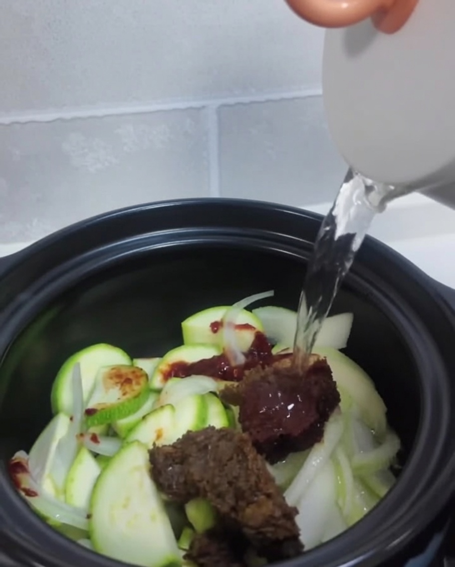 Klares Wasser wird aus einem weißen Wasserkocher über das Gemüse und die Pasten in den Topf gegossen.