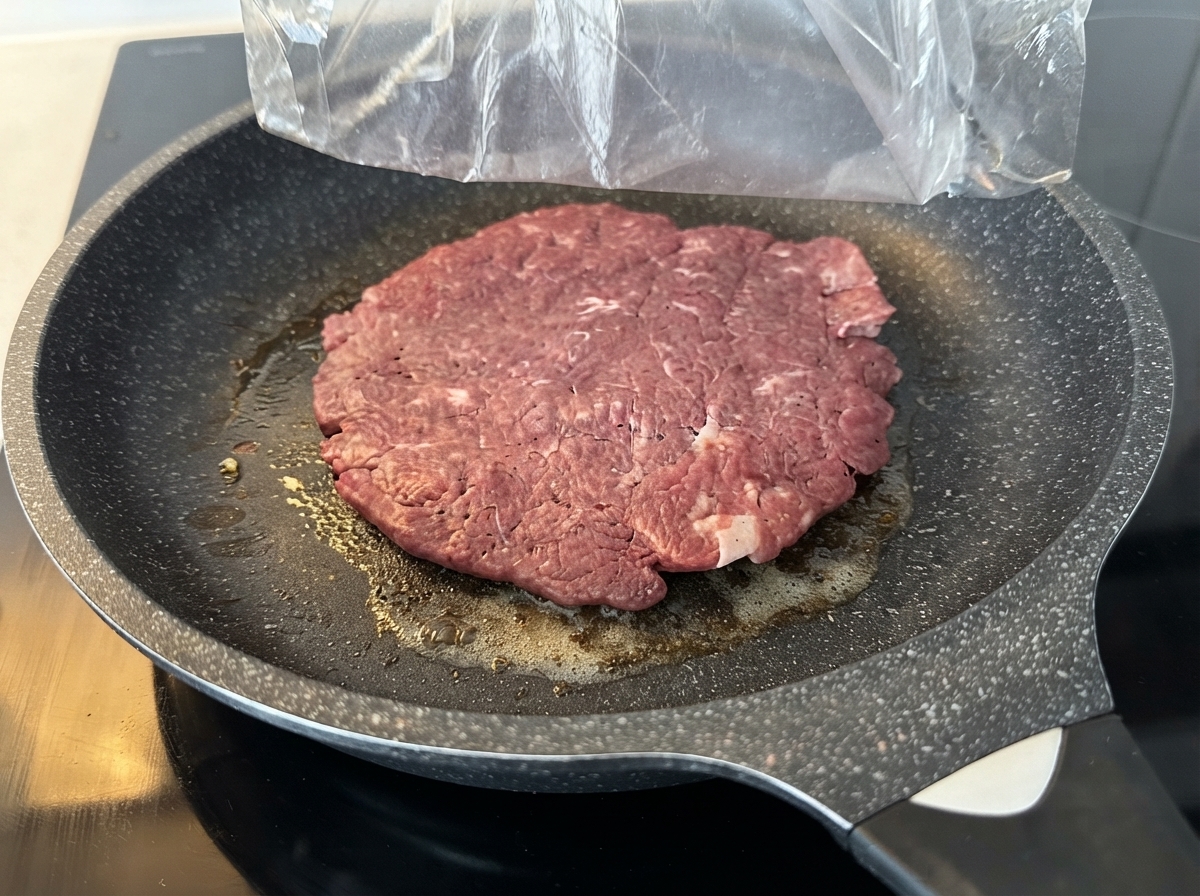 Ein großes rohes Rinderpatties brät in einer gefetteten Antihaft-Pfanne auf einem Herd.