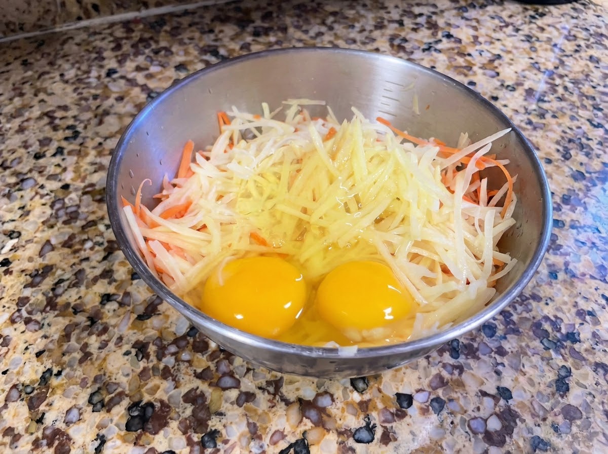 Eine Metallschüssel mit frisch geriebenen Kartoffeln und Karotten sowie zwei ganzen Eiern darauf.