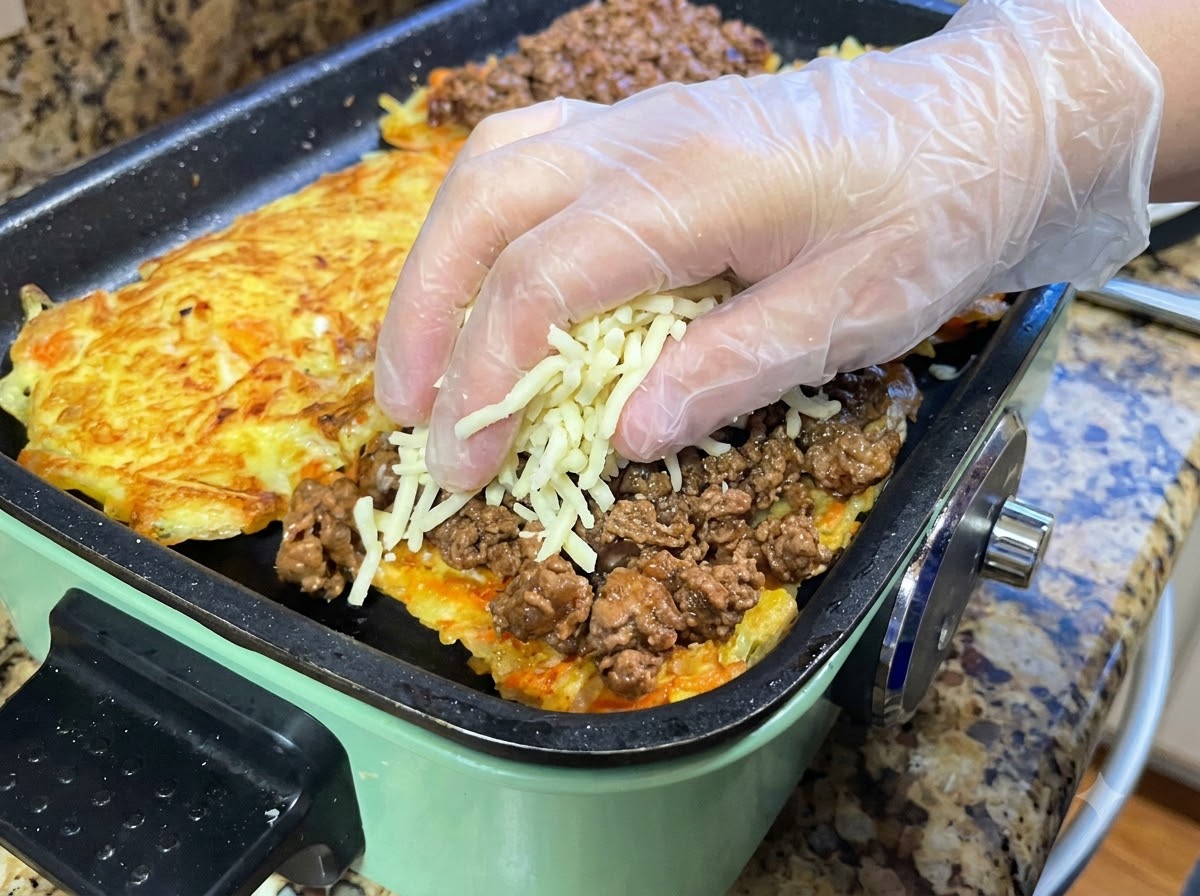 Hände in Schutzhandschuhen streuen eine Handvoll geriebenen Mozzarella über eine Schicht Fleischsauce auf einem Kartoffelrösti.