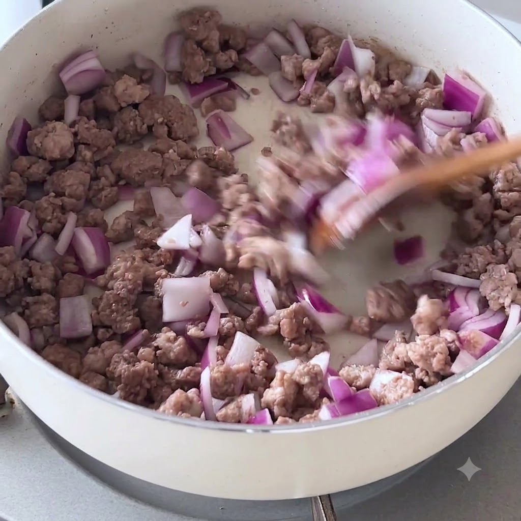 Gehackte rote Zwiebeln und angebratenes Hackfleisch werden mit einem Holzwender in einer weißen Pfanne verrührt.
