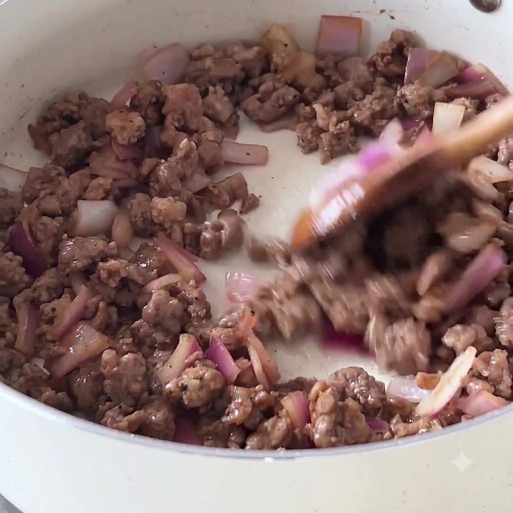 Anbraten von Hackfleisch und roten Zwiebeln in einer weißen Antihaft-Pfanne mit einem Holzlöffel.