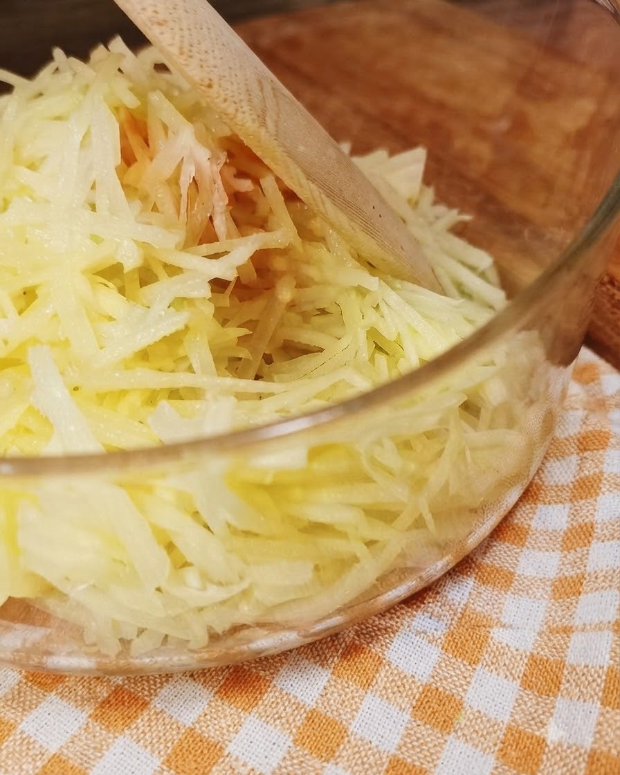 Frisch geriebene Kartoffeln in einer klaren GlasSchüssel mit einem Holzlöffel.