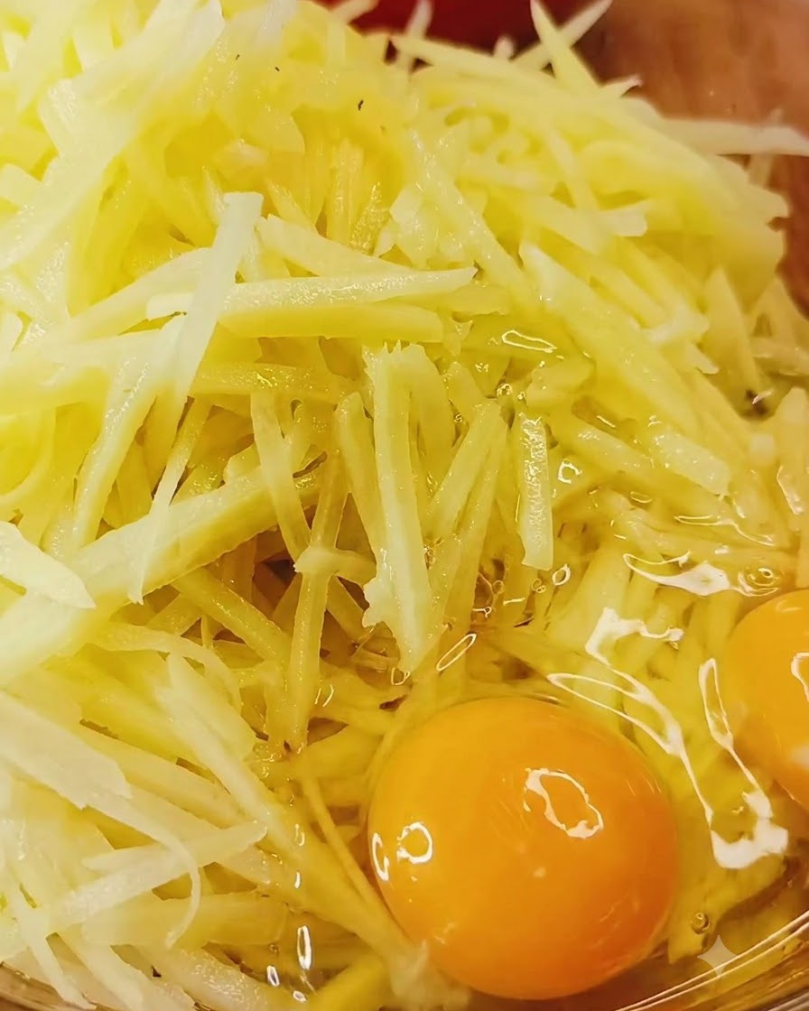 Ganze Eier direkt über einer Schüssel mit rohen Kartoffelraspeln und Gewürzen aufgeschlagen.