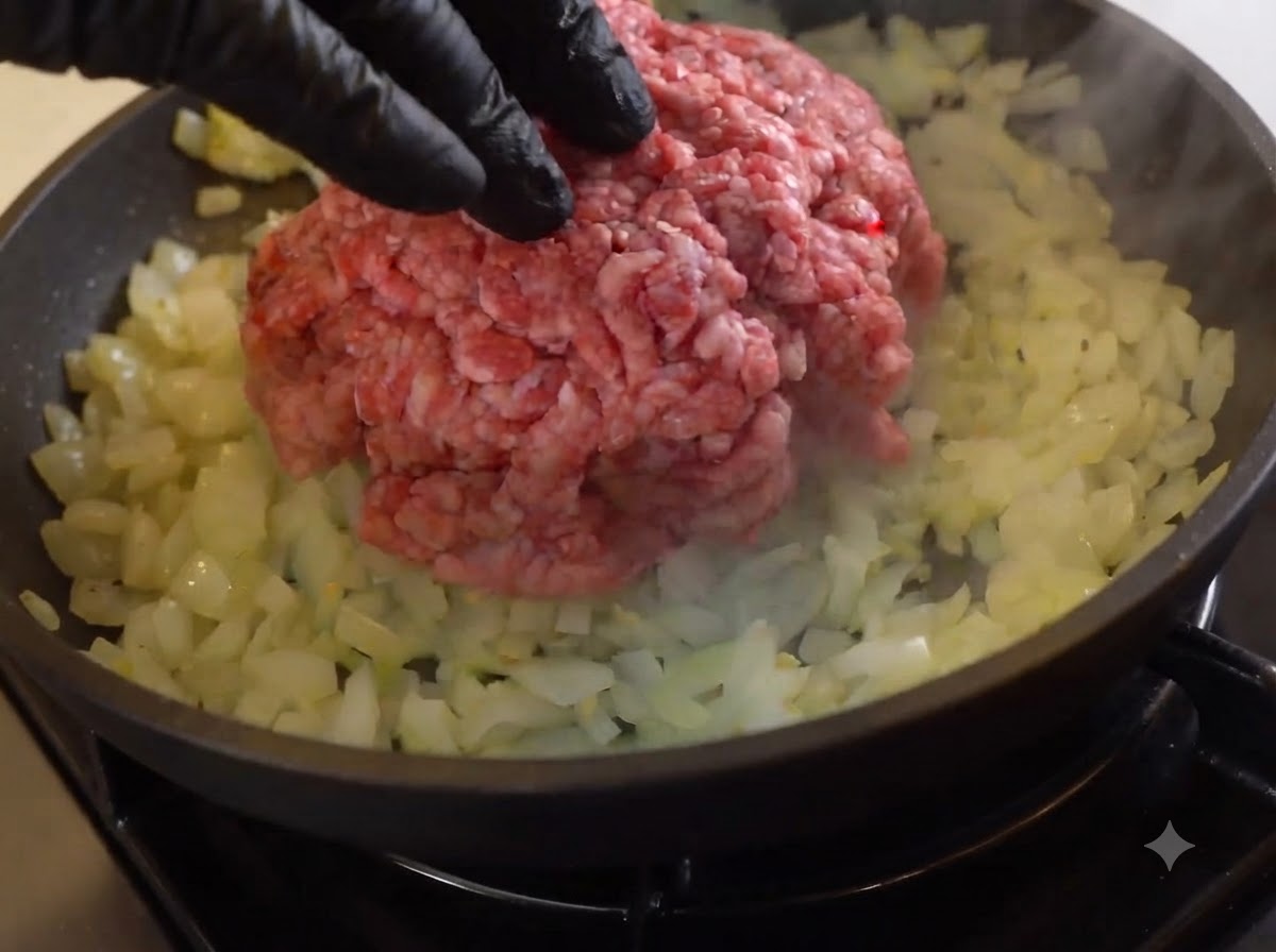Ein Berg rohes Rinderhackfleisch liegt auf angebratenen Zwiebeln in einer Pfanne.