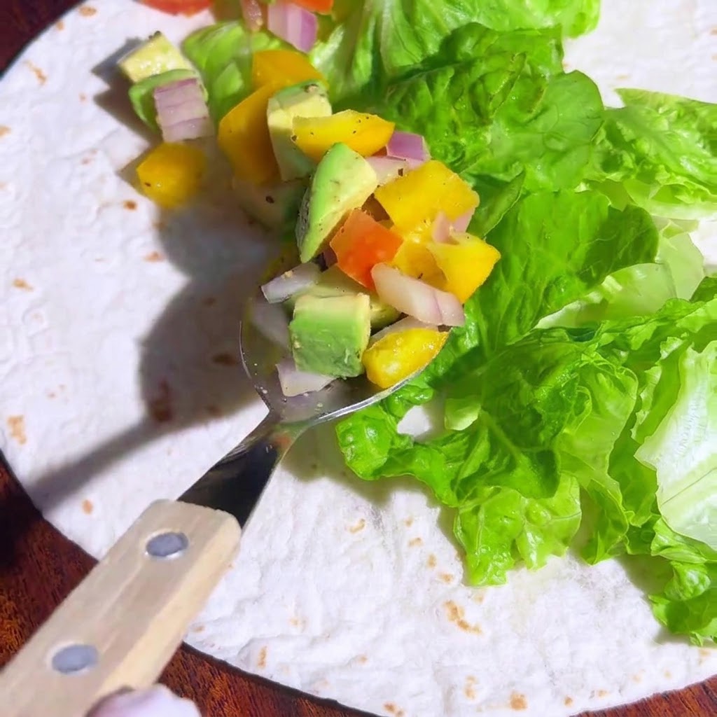 Ein Löffel mit bunter gewürfelter Avocado-Gemüse-Salsa, der über dem Salat auf einer Tortilla positioniert ist.