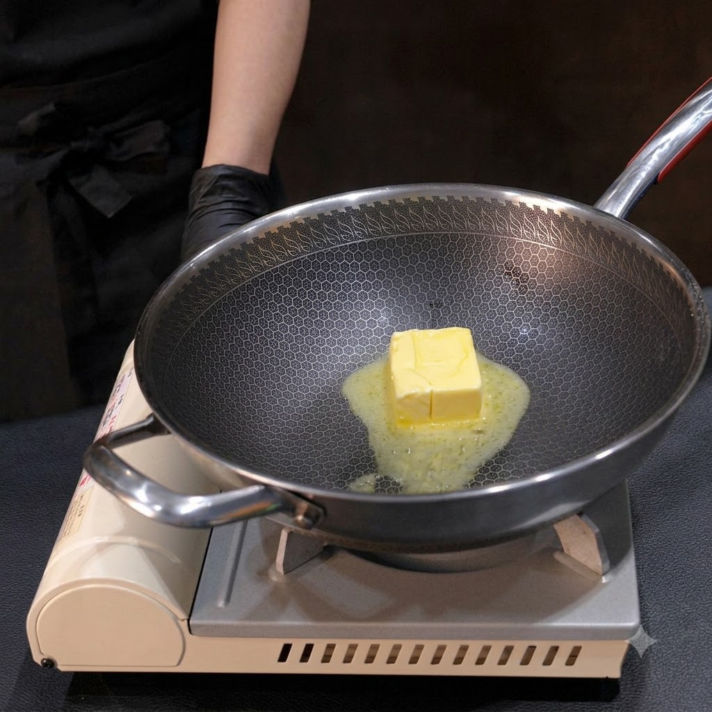 Ein Block gelbe Butter schmilzt in einem Metall-Wok und wird mit einem kleinen Schneebesen über Hitze gerührt.