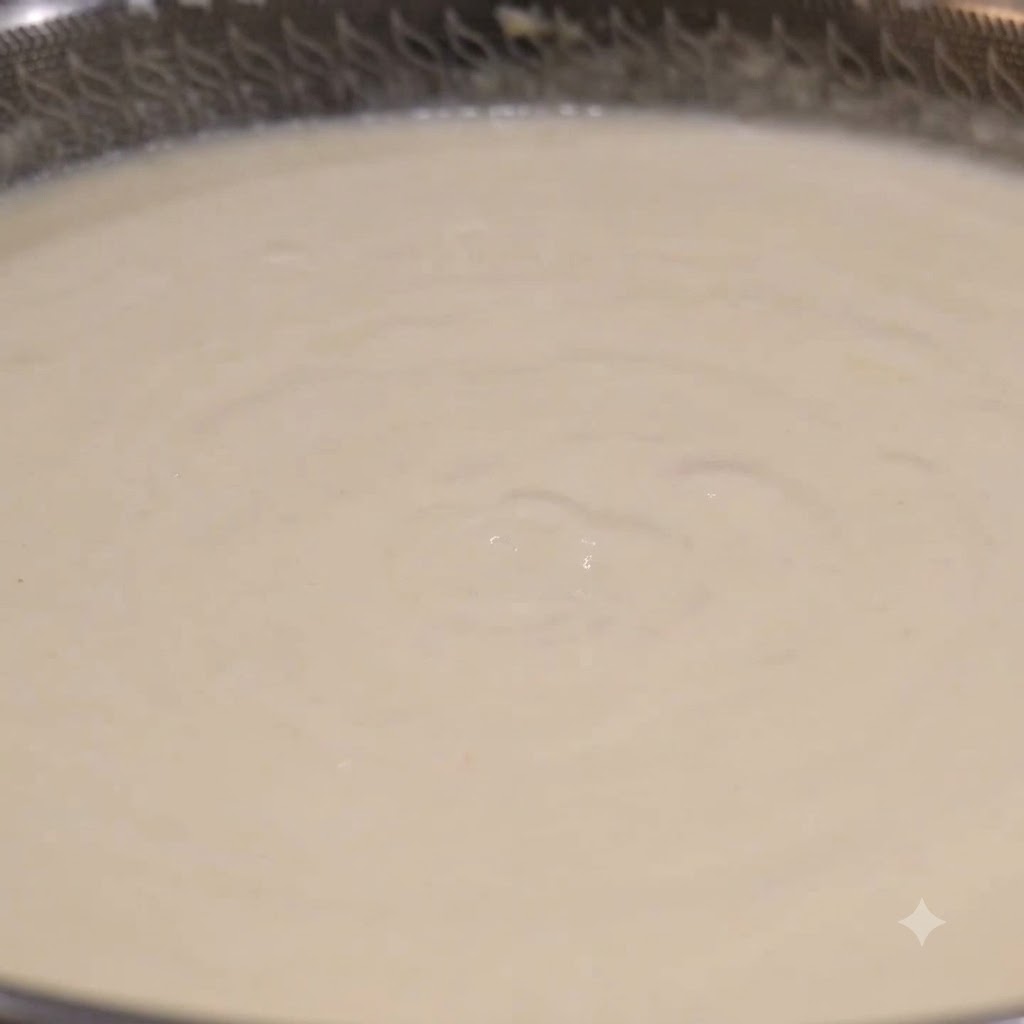 Eine Schöpfkelle schöpft cremige weiße Muschelsuppe aus einem köchelnden Wok.
