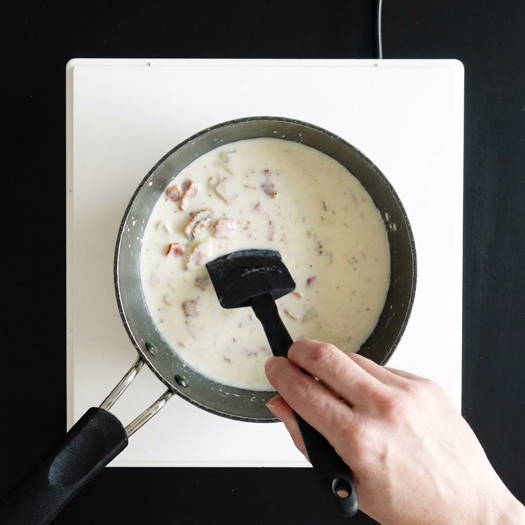 Eine Hand rührt eine dicke, cremige weiße Suppe in einem Stieltopf mit einem schwarzen Silikonspatel um.