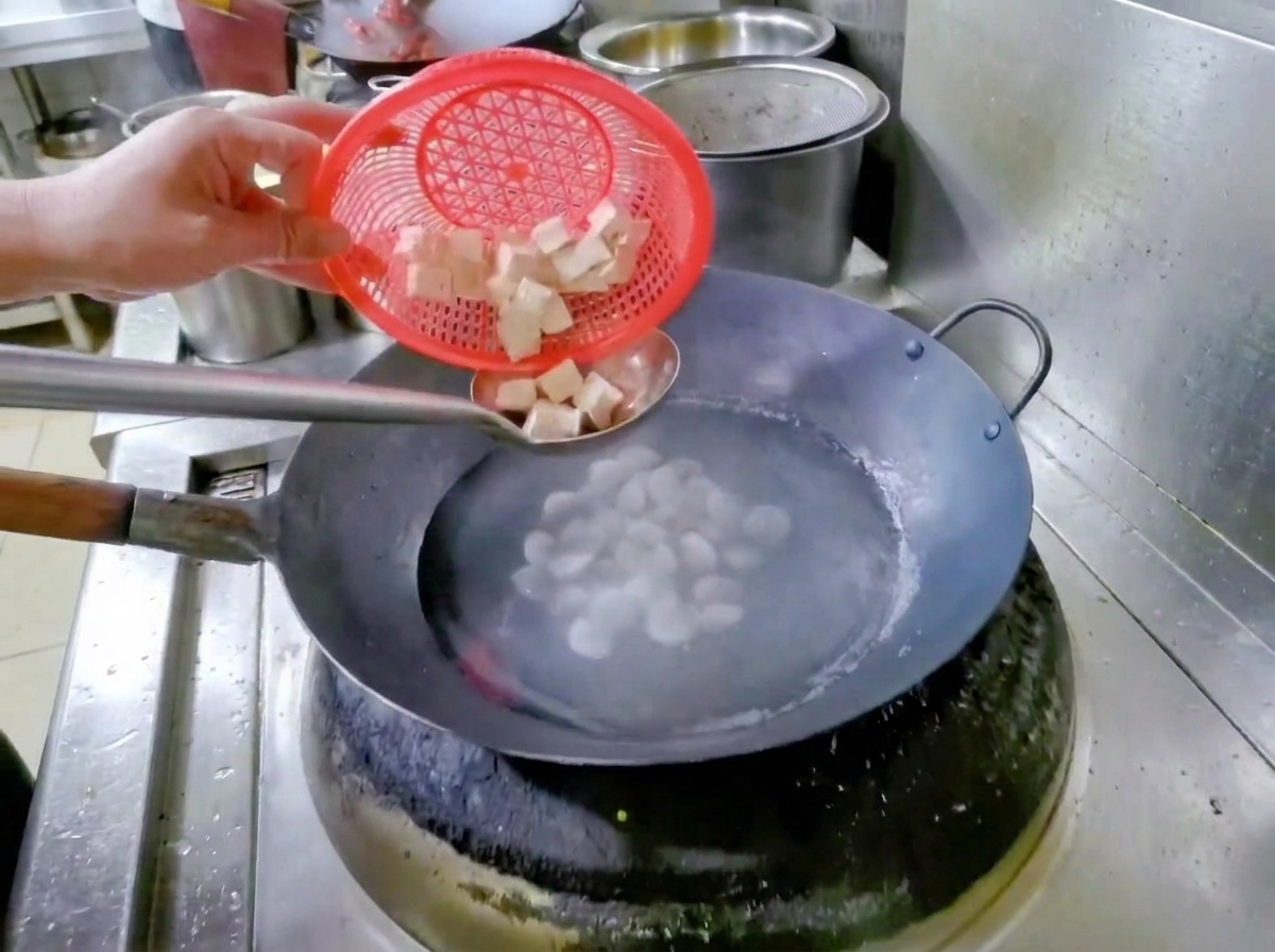 Eine Hand hält ein rotes Sieb und gibt gewürfelten Tofu in einen Wok mit Wasser und Muscheln.