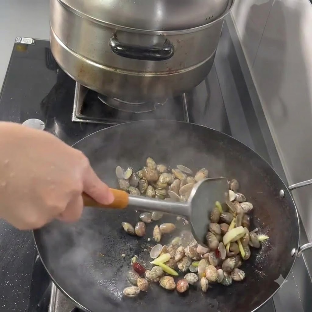 Muscheln, Knoblauch, Ingwer und Chilis werden mit einem Metallspatel in einem Wok angebraten.