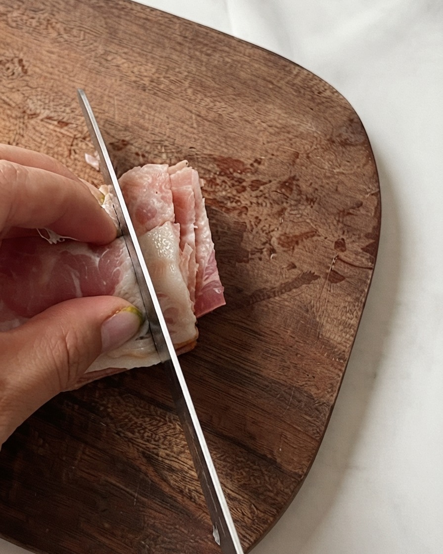 Eine Hand hält gestapelte Speckstreifen, während ein Messer sie auf einem Holzbrett in kleine Stücke schneidet.