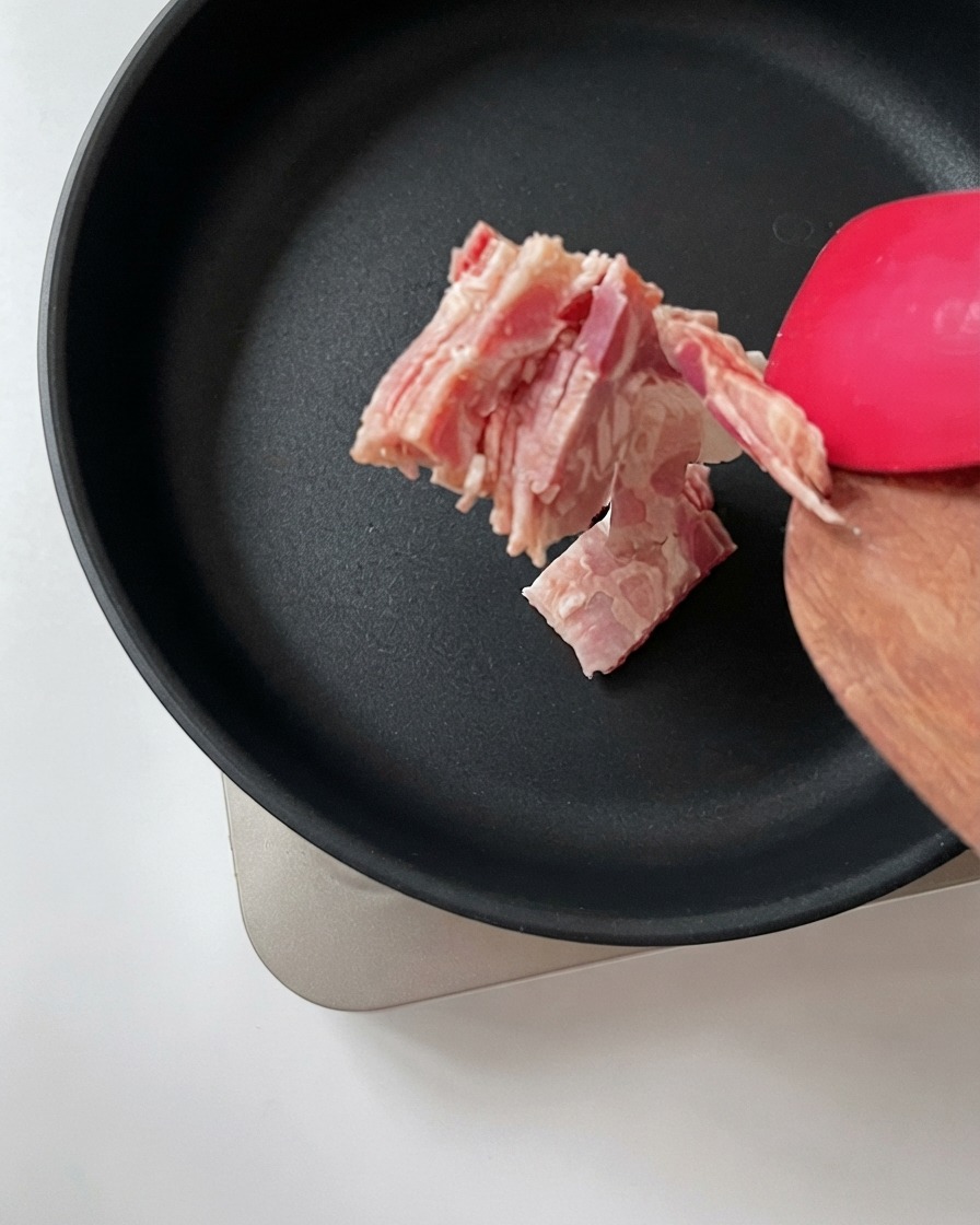 Gehackter Speck wird mit einem Holzspatel in eine heiße schwarze Bratpfanne gegeben.