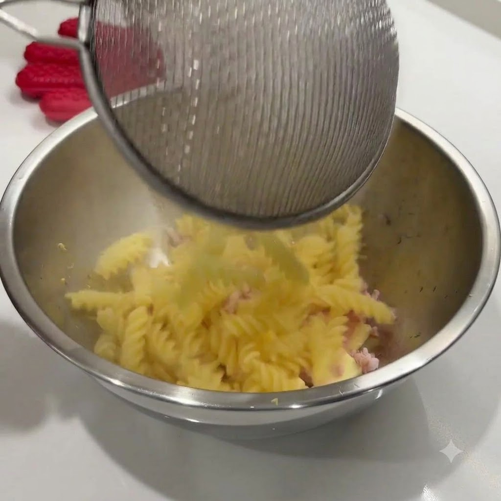 Gekochte Spiralnudeln werden aus einem Drahtsieb in eine Edelstahlschüssel mit den Zutaten geschüttet.