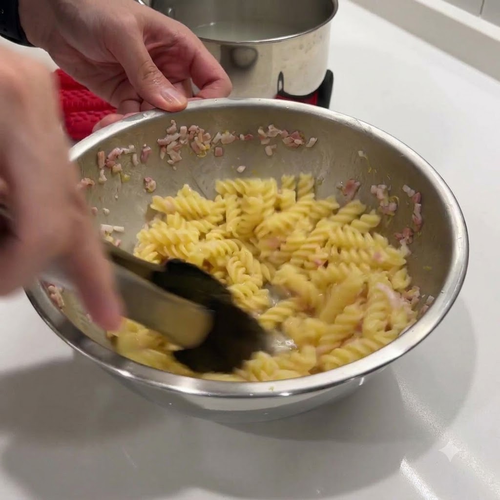 Heisse Spiralnudeln und Butter werden in einer Metallschüssel mit einer schwarzen Zange umgerührt.