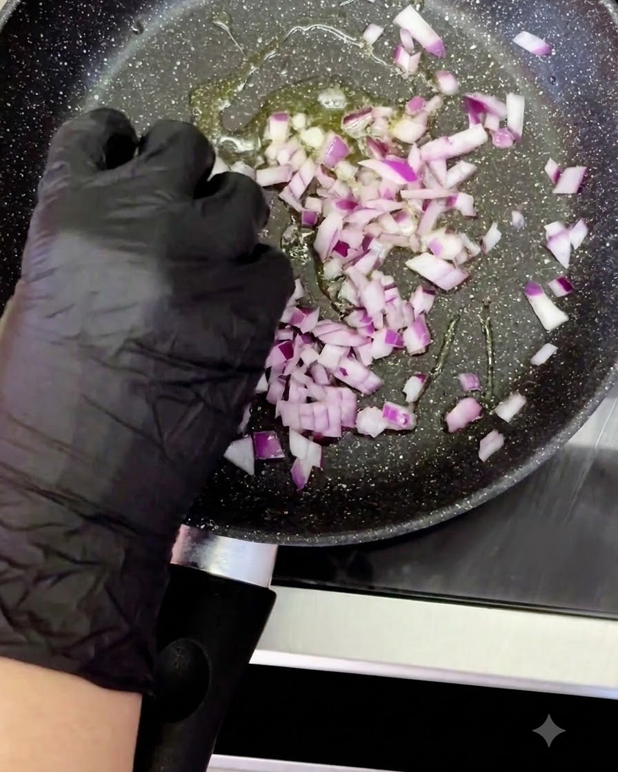 Eine behandschuhte Hand gibt gewürfelte rote Zwiebeln in eine erhitzte Pfanne mit brutzelndem Öl.